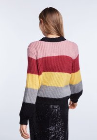Donna di spalle che indossa un maglione lavorato a maglia con larghe righe orizzontali nei colori rosa, rosso, giallo, grigio e nero, abbinato a una gonna nera con paillettes.