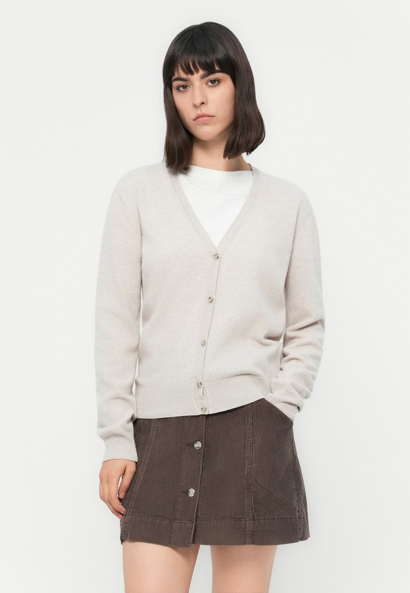 Una giovane donna con capelli corti e scuri indossa un cardigan beige chiaro con bottoni sopra una maglietta bianca e una gonna in velluto corduroy marrone scuro con bottoni.