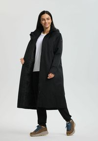 Kobieta w czarnym długim płaszczu, białej koszuli, czarnych spodniach i brązowych butach górskich z niebieskimi sznurówkami, stojąca i uśmiechająca się na jednolitym tle.