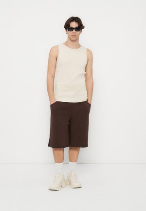 Ung mann iført beige ermeløs skjorte, mørkebrune knelang shorts, hvite sokker, kraftige joggesko og svarte solbriller, stående med hendene i lommer.