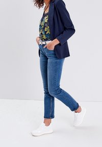 Blazer bleu marine sur un caraco à fleurs, associé à un jean bleu et des baskets blanches. L'ensemble comprend une ceinture et un tissu texturé.