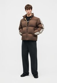 Junger Mann, der eine braune Steppjacke mit weißen Streifen an den Ärmeln, schwarze Hose und schwarze Schuhe trägt, vor einem schlichten weißen Hintergrund steht.