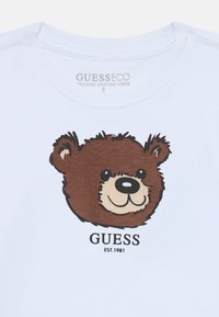 Weißes T-Shirt aus Bio-Baumwolle mit einem braunen Cartoon-Bärenkopf und dem Aufdruck "GUESS EST. 1981" darunter in Schwarz.