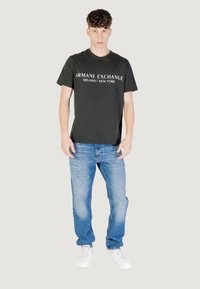 T-shirt in cotone grigio scuro con logo "Armani Exchange" in bianco, abbinata a jeans in denim azzurro chiaro e sneakers bianche. Sfondo semplice.