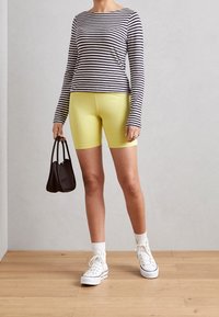 Top a maniche lunghe a righe blu navy e bianche, abbinato a pantaloni corti gialli aderenti. Gli accessori includono una piccola borsa nera e sneakers bianche.