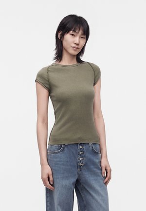 Jeune femme aux cheveux noirs raides portant un haut ajusté manches courtes vert olive et un jean bleu taille haute avec des boutons.