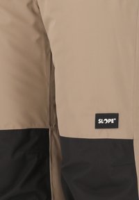 Pantaloni da sci beige e neri, caratterizzati da una texture liscia, blocchi di colore a contrasto e un'etichetta ricamata sul lato con la scritta "SLOPE."