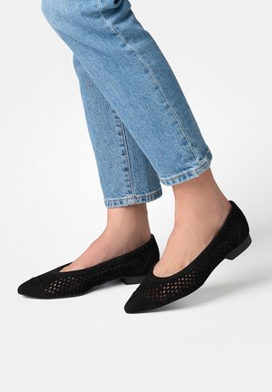 Beine in hellblauen Jeans, die schwarze spitze Ballerinas mit perforiertem Mesh-Design und durchgehenden Zehenkappen vor weißem Hintergrund tragen.