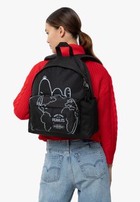 Mochila negra con un gran bolsillo frontal, diseño de dibujo animado contorneado y detalle de logo; llevada por un modelo con un suéter rojo.