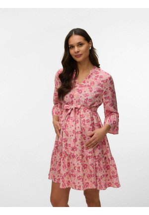 Ženska z dolgimi temnimi lasmi, oblečena v rožnato cvetlično obleko do kolen, drži pas in stoji pred enobarvnim belim ozadjem.