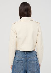 Giacca corta di un beige chiaro realizzata in materiale sintetico liscio, con colletto a punte e spalline. Dettaglio della cucitura inclinata sulla schiena.