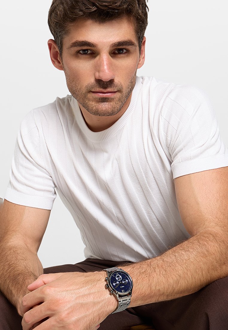 Reloj plateado con una esfera azul redonda, pulsera de acero inoxidable, llevado en una muñeca. Modelo con una camiseta blanca de manga corta con canalé, sentado.