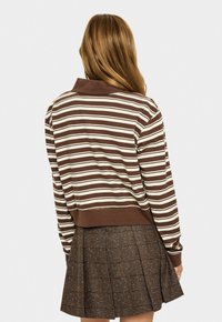 Femme avec de longs cheveux châtain clair portant un haut à rayures marron et blanc à manches longues et une jupe plissée écossaise marron, de dos.