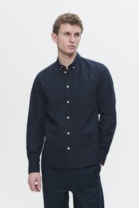Young man wearing a fitted dark navy buttoned shirt with long sleeves and a chest pocket, standing against a plain light background.