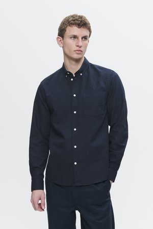 Young man wearing a fitted dark navy buttoned shirt with long sleeves and a chest pocket, standing against a plain light background.