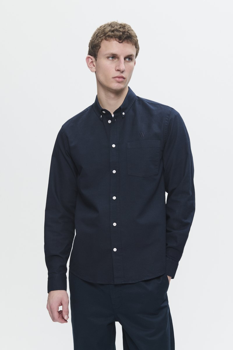 Young man wearing a fitted dark navy buttoned shirt with long sleeves and a chest pocket, standing against a plain light background.