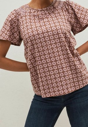 Kobieta ubrana w różowo-brązową krótki rękaw bluzkę w kwiaty z marszczonym karczkiem, w połączeniu z ciemnoniebieskimi dżinsami, stoi z rękami na biodrach.