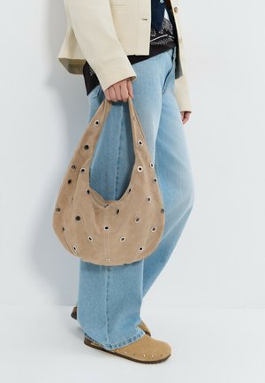 Persona vistiendo vaqueros azul claro, chaqueta beige y zapatos de ante beige, sosteniendo un bolso de ante beige con detalles de ojales metálicos.