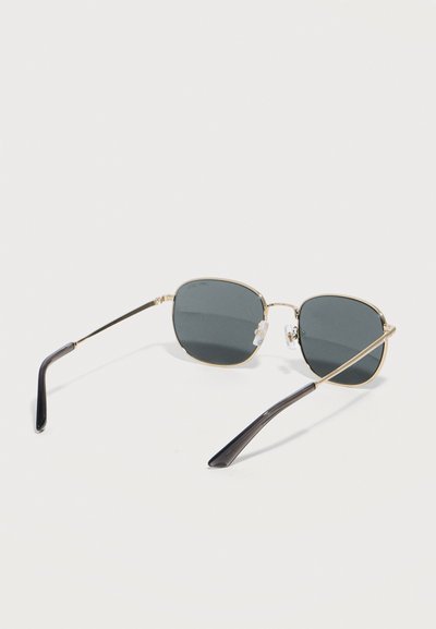 Κομψά γυαλιά ηλίου με λεπτά χρυσά τετράγωνα πλαίσια, σκοτεινούς φακούς, μαύρες καμπυλωτές άκρες στο ναύσταθμο και ρυθμιζόμενα μαξιλαράκια μύτης σε λευκό φόντο.