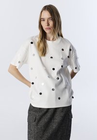 Weißes kurzärmeliges Hemd mit runden metallischen Verzierungen in verschiedenen Größen, locker geschnitten und mit rundem Ausschnitt. Kombiniert mit strukturierten grauen Hosen.