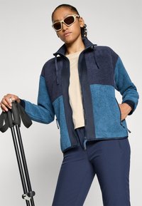 Person med beige topp, blå og marinblå fleecejakke, mørkeblå bukser og hvite solbriller, som holder svarte trekkingstaver mot en lys bakgrunn.