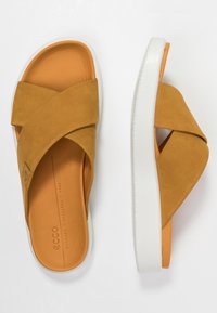 Des sandales à glissière en suède marron avec des bandes croisées, doublées d'orange à l'intérieur et dotées d'une semelle en caoutchouc blanche. Elles disposent d'une semelle intérieure rembourrée et d'un design minimaliste.