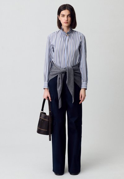 Camisa de botón a rayas con cuello alto, suéter gris atado a la cintura y pantalones cargo azul marino. Bolso negro con detalles dorados.
