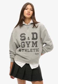 Grijze sweatshirt met grote zwarte letters "S&D GYM ATHLETIC" en geborduurd logo, gelaagd over een witte coll shirt, gecombineerd met een zwarte geplooide rok.