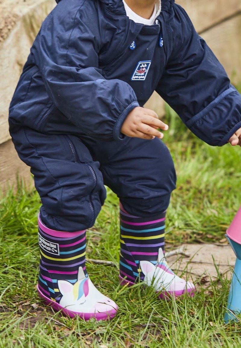 Enfant portant une combinaison de pluie bleu marine et des bottes de pluie rayées avec des visages de licorne, debout sur l'herbe près d'une surface en bois et d'un objet bleu.