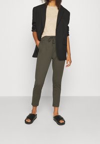 Donna che indossa un blazer nero, un maglione beige, pantaloni verde oliva con coulisse e sandali neri, mentre si trova davanti a uno sfondo bianco semplice.
