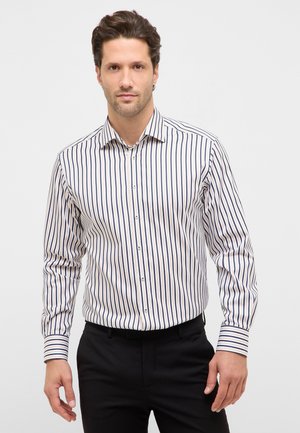 Mand iført en langærmet, hvid skjorte med knapper og lodrette sorte og brune striber, kombineret med sorte bukser.