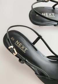 Schwarze Patentleder-Sandalen mit schlanken, verstellbaren Riemen und einer markierten Innensohle. Sie verfügen über eine abgerundete Zehenpartie und goldfarbene Hardware-Akzente.