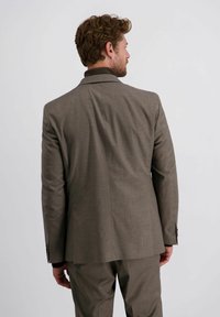 Mann mit lockigem braunem Haar, der einen braunen Anzug und einen dunklen Rollkragenpullover trägt und mit dem Rücken zu einem schlichten hellen Hintergrund steht.