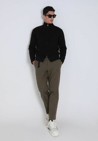 Pull à zip noir côtelé, pantalon olive avec un nœud sur le côté, et baskets blanches. Modèle portant des lunettes de soleil noires, posant sur un fond clair.