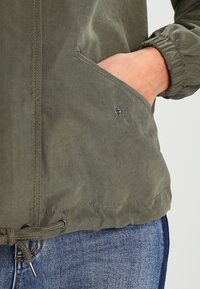 Chaqueta verde oliva con puños elásticos, dobladillo ajustable y un bolsillo lateral con un detalle de botón a presión. Fabricado en un tejido suave y texturizado, de diseño casual.