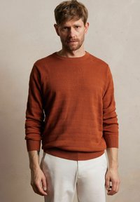 Spalony pomarańczowy sweter dzianinowy z teksturowanymi paskami, okr�ągłym dekoltem i ściągaczami przy rękawach. Zestawiony z jasnymi spodniami.