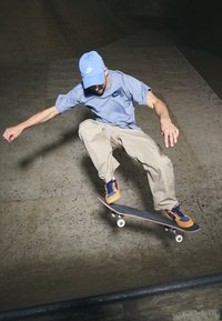 Rullalautaaja sinisessä Nike SB -t-paidassa ja lippiksessä, joka tekee temppua rullalaudalla, jossa on oranssit ja laivastonsiniset sneakerit, päällään vaaleanbeige housut, skeittipuistossa.