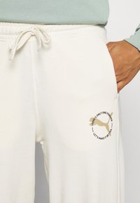 Cream-colored sweatpants with a soft texture, featuring a gold logo and text design on the left thigh. Elastic waistband with adjustable drawstring.