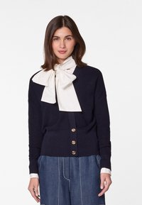 Marineblauer Strickcardigan mit goldenen Knöpfen und einem dezenten V-Ausschnitt. Kombiniert mit einem cremefarbenen Hemd mit Schleifenkragen und lockeren Jeanshosen.