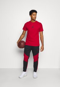 Rød T-shirt med korte ærmer fra Jordan med sort logo, parret med sorte joggers med røde detaljer og hvide sportssko, mens han holder en basketball.
