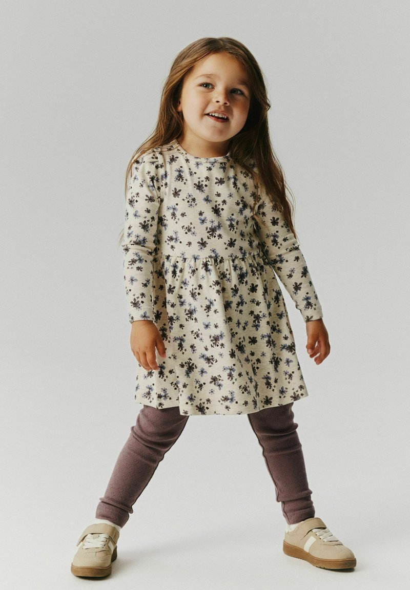 Jeune fille debout, jambes écartées, portant une robe fleurie à manches longues, des leggings violets et des baskets beiges, souriante et regardant légèrement vers la droite.