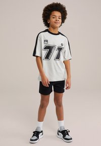 Svetlo siva majica s kratkimi rokavi z črnimi detajli, ki vsebuje velik črn številko "71" in logo. V kombinaciji z črnimi hlačami in športnimi copati.