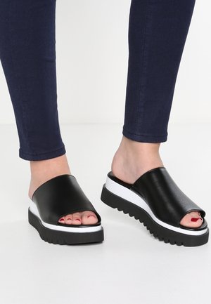 Pieds chaussés de sandales plateformes noires à enfiler avec des semelles intermédiaires blanches et des semelles extérieures noires dentelées, portées avec un pantalon skinny bleu foncé.