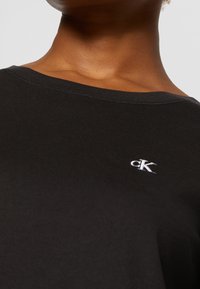 Detailní záběr na osobu, která má na sobě černou košili s malým bílým vyšívaným logem "cK" poblíž výstřihu.