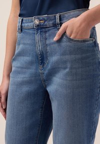 Vaqueros de mezclilla azul claro con un diseño ajustado, que cuentan con un botón decorativo plateado, un bolsillo frontal y una textura de desvanecimiento sutil.
