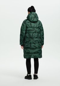 Abrigo de plumas verde de camuflaje con capucha, que presenta una textura acolchada y un diseño de longitud media, combinado con leggings y zapatos negros.