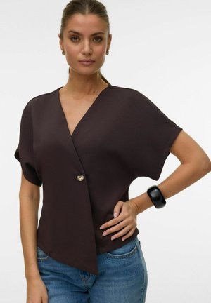 Kobieta z zaczesanymi do tyłu włosami, ubrana w ciemny top w stylu kopertowym z pojedynczym złotym guzikiem, niebieskie jeansy i masywną czarną bransoletkę.