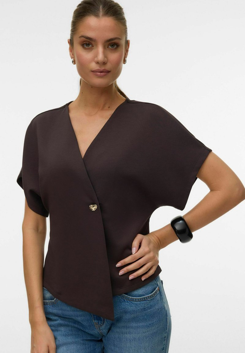 Kobieta z zaczesanymi do tyłu włosami, ubrana w ciemny top w stylu kopertowym z pojedynczym złotym guzikiem, niebieskie jeansy i masywną czarną bransoletkę.