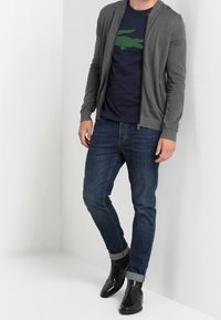 Cardigan grigio con zip sopra una t-shirt blu navy con logo di coccodrillo verde, abbinato a jeans blu e stivali alla caviglia neri.