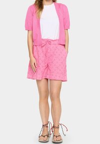 Vaaleanpunainen, lyhythihainen neuletakki ja siihen sopivat nyörillä varustetut Shortsit, joissa on brodeerattuja kukkakuvioita. Jalkineissa on sekoitettujen värien strapit.